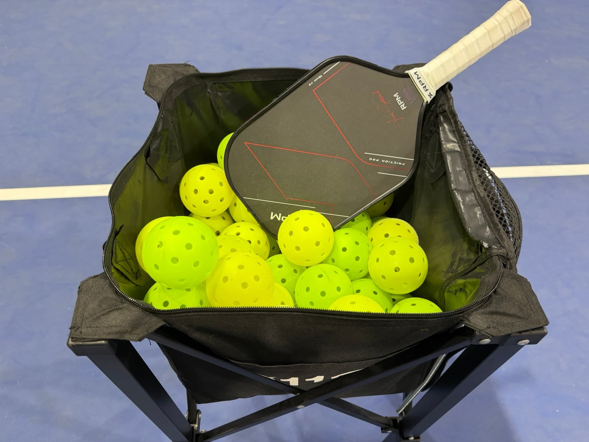 An RPM paddle resting on a hopper full of pickleballs